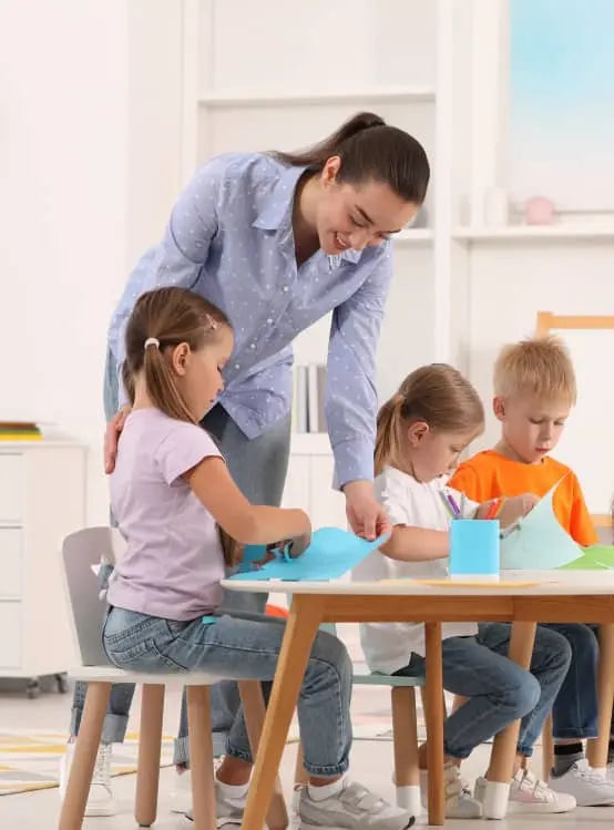 Nursery teacher with group of cute little children drawing and cutting paper at desks in kindergarten